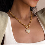 Load image into Gallery viewer, A close-up of a woman wearing a gold chain necklace with a toggle clasp and a textured heart-shaped pendant, styled with a white ribbed tank top and a light olive green cardigan.