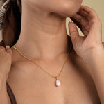 Load image into Gallery viewer, A close-up of a woman's neck and chest as she wears a delicate gold snake chain necklace with a single baroque pearl pendant. Her hand gently touches the side of her neck, and she is wearing a brown patterned headscarf.