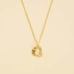 Load image into Gallery viewer, A clean, minimalist product shot of a modern gold and pearl necklace on a neutral cream background. The image clearly displays the sculptural, ribbed gold cage pendant design with a single, lustrous pearl nestled inside.