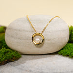 Load image into Gallery viewer, A contemporary gold necklace from Zewory Jewellery, featuring an open circle pendant with a central pearl. The elegant, minimalist piece is displayed on a smooth, light-grey stone and surrounded by fresh green moss for a natural aesthetic.