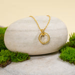 Load image into Gallery viewer, An elegant gold circle necklace from Zewory Jewellery, displayed in a serene natural setting. The sparkling pavƩ crystal and twisted rope-textured pendant is draped over a smooth, light-grey stone and surrounded by fresh green moss.
