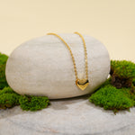 Load image into Gallery viewer, A delicate gold heart necklace from Zewory Jewellery, displayed in a serene natural setting. The minimalist, polished heart pendant is draped over a smooth, light-grey stone and surrounded by fresh green moss.