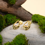 Load image into Gallery viewer, A pair of contemporary gold and pearl stud earrings displayed in a natural setting. The organic, open-frame design of each earring holds a single pearl, beautifully showcased on a grey stone with moss and weathered wood.