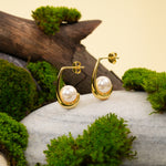 Load image into Gallery viewer, A pair of contemporary gold and pearl J-hook earrings, displayed in a natural setting. The modern design with a cradled pearl in each earring is showcased on a grey stone, surrounded by vibrant green moss and weathered wood.