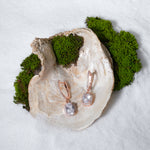 Load image into Gallery viewer, A luxurious product shot of sparkling rose gold halo drop earrings. This elegant, bridal-style pair is artistically arranged inside a large seashell on a bed of white sand and moss.