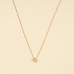 Load image into Gallery viewer, A clean, minimalist product shot of a rose gold solitaire necklace on a neutral cream background. The image clearly displays the delicate chain and the classic, round brilliant-cut stone in a modern bezel setting.