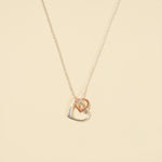 Load image into Gallery viewer, A clean, minimalist product shot of a two-tone interlocking hearts necklace on a neutral cream background. The image clearly displays the delicate silver chain with its mixed-metal, double-heart pendant.
