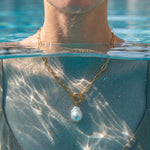 Load image into Gallery viewer, A gold chain necklace with a toggle clasp and a single baroque pearl pendant, worn by a person in shallow water. The image is split horizontally by the waterline, with the person's neck and upper chest visible above the water and the necklace hanging below.