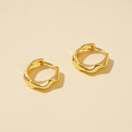 Load image into Gallery viewer, A clean product shot of a pair of minimalist gold huggie hoops with a modern, irregular wavy design. The image clearly displays the sculptural shape and high-polish finish of the earrings against a neutral cream background.