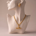 Load image into Gallery viewer, A gold necklace with a bar pendant displayed on a white textured mannequin bust, with a matching gold seashell earring shown on the mannequin's ear against a warm beige background.
