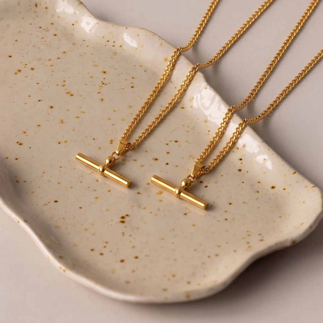 Two gold chain necklaces with minimalist bar pendants resting on a speckled cream ceramic dish.