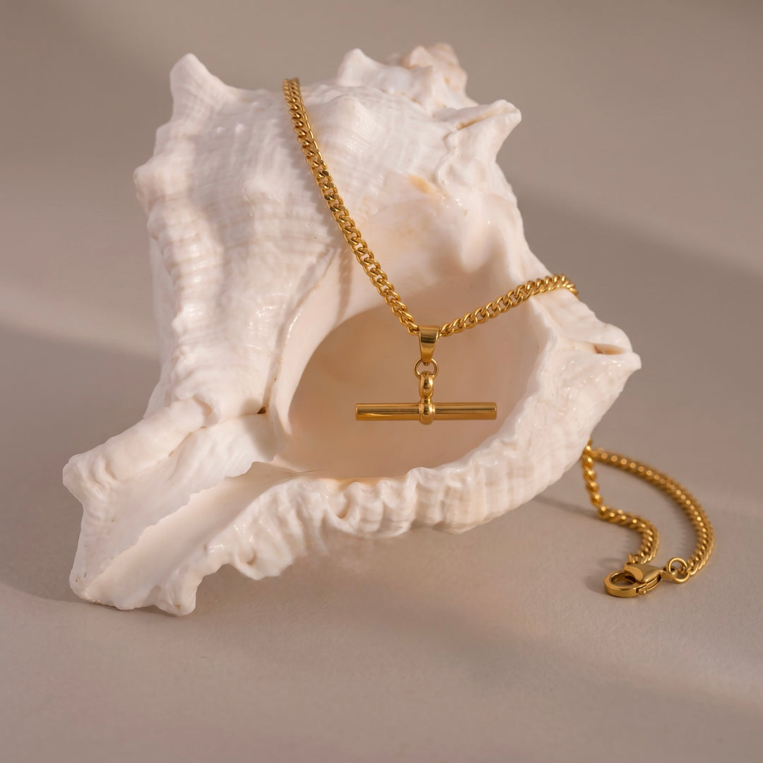 A gold chain necklace with a toggle clasp pendant, draped over a large white seashell on a neutral beige background.