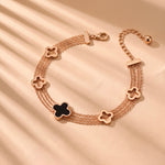 Load image into Gallery viewer, A rose gold multi-strand chain bracelet with five clover-shaped charms, four in rose gold and one in black enamel, displayed on a beige fabric background with soft shadows.

