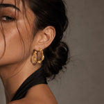 Load image into Gallery viewer, A close-up of a woman's ear wearing a textured, twisted gold hoop earring against a neutral gray background.
