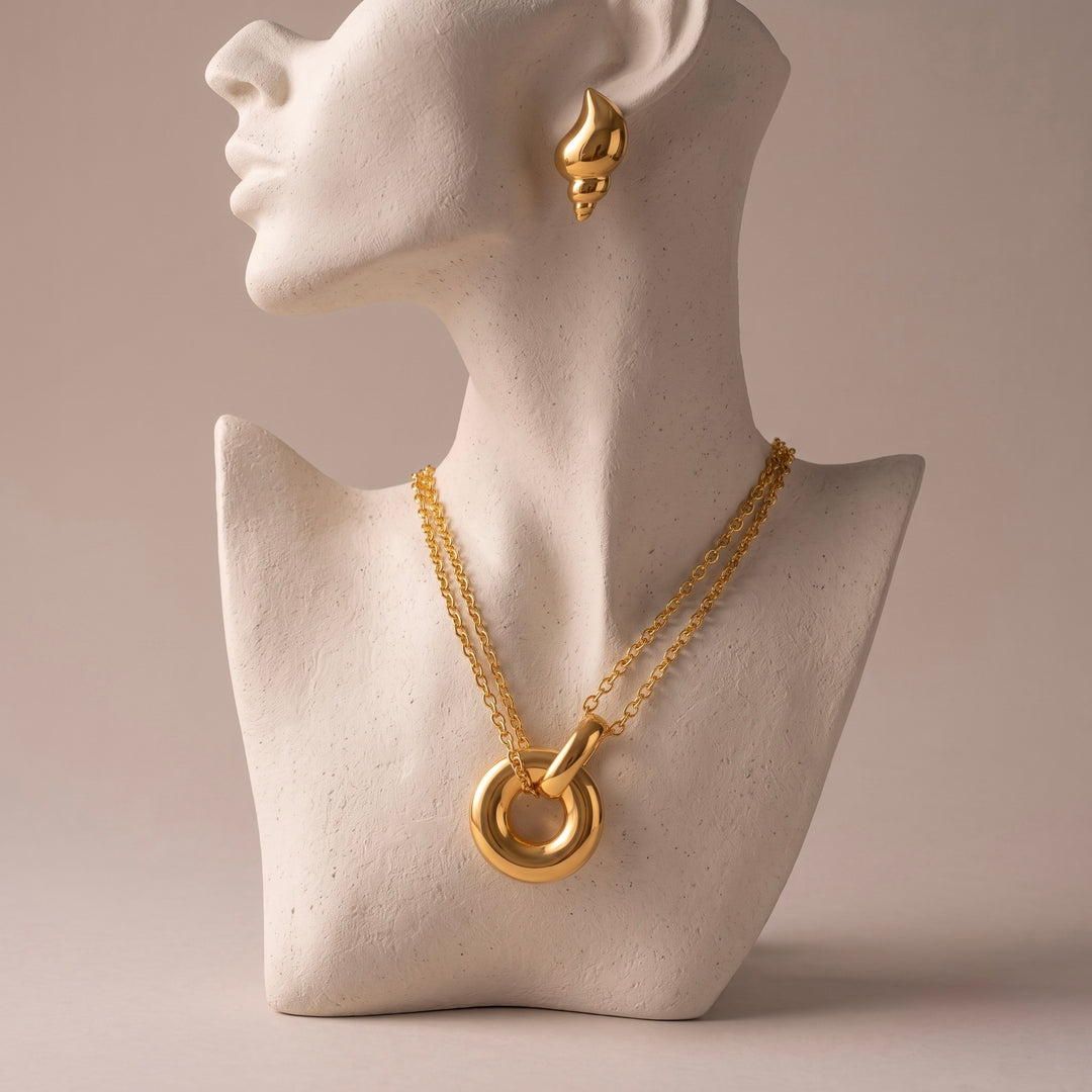 A gold necklace with a large interlocking ring pendant displayed on a white textured bust, with a matching gold teardrop earring shown on the bust's ear against a warm beige background.