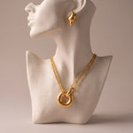 Load image into Gallery viewer, A gold necklace with a large interlocking ring pendant displayed on a white textured bust, with a matching gold teardrop earring shown on the bust's ear against a warm beige background.
