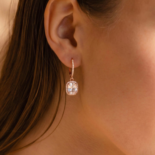 Close-up of a model wearing rose gold drop hoop earrings with rectangular clear stones and sparkling halo accents.