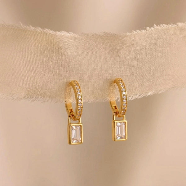Gold hoop earrings with rectangular gemstones on a beige fabric background