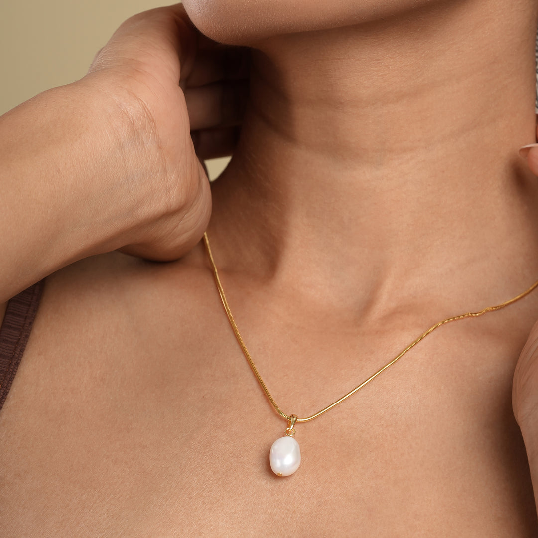 A close-up of a woman's neck and chest as she wears a delicate gold snake chain necklace with a single baroque pearl pendant. Her hand gently touches the side of her neck, and she is wearing a brown patterned headscarf.