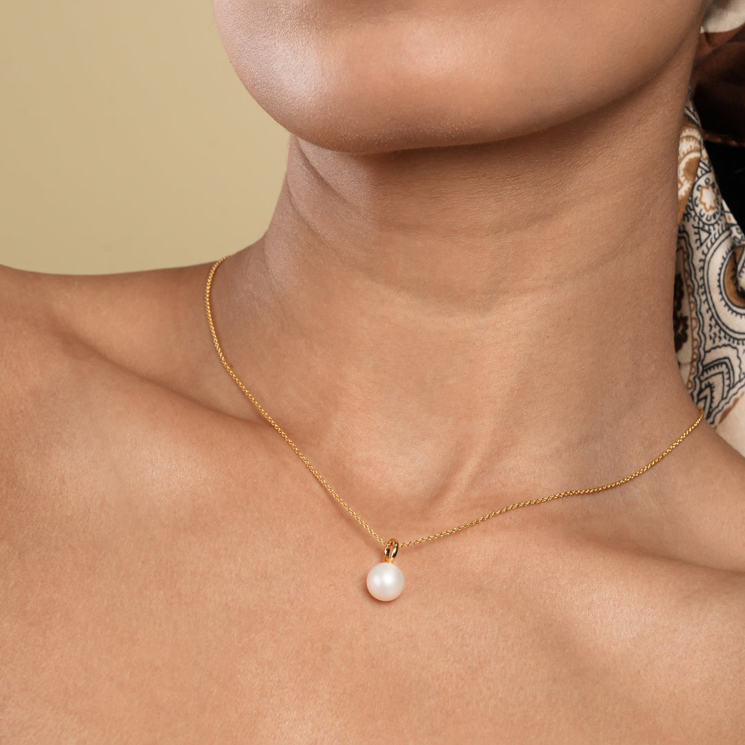 A close-up of a woman's neck and chest as she wears a delicate gold snake chain necklace with a single baroque pearl pendant. Her hand gently touches the side of her neck, and she is wearing a brown patterned headscarf.