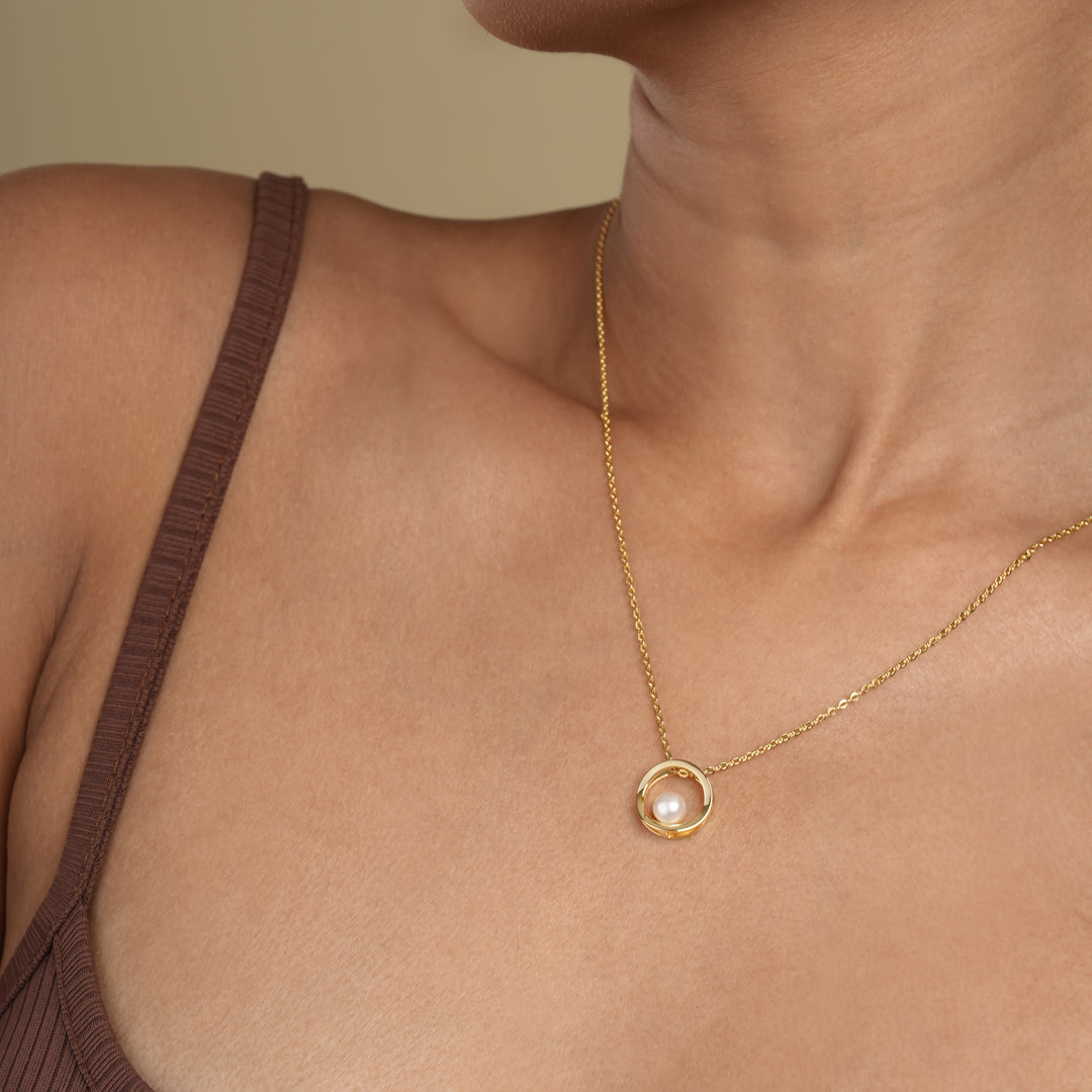 A close-up of a model wearing a modern gold and pearl circle necklace from Zewory Jewellery. The elegant pendant, featuring a single pearl suspended within a polished gold hoop, rests gracefully on her chest.