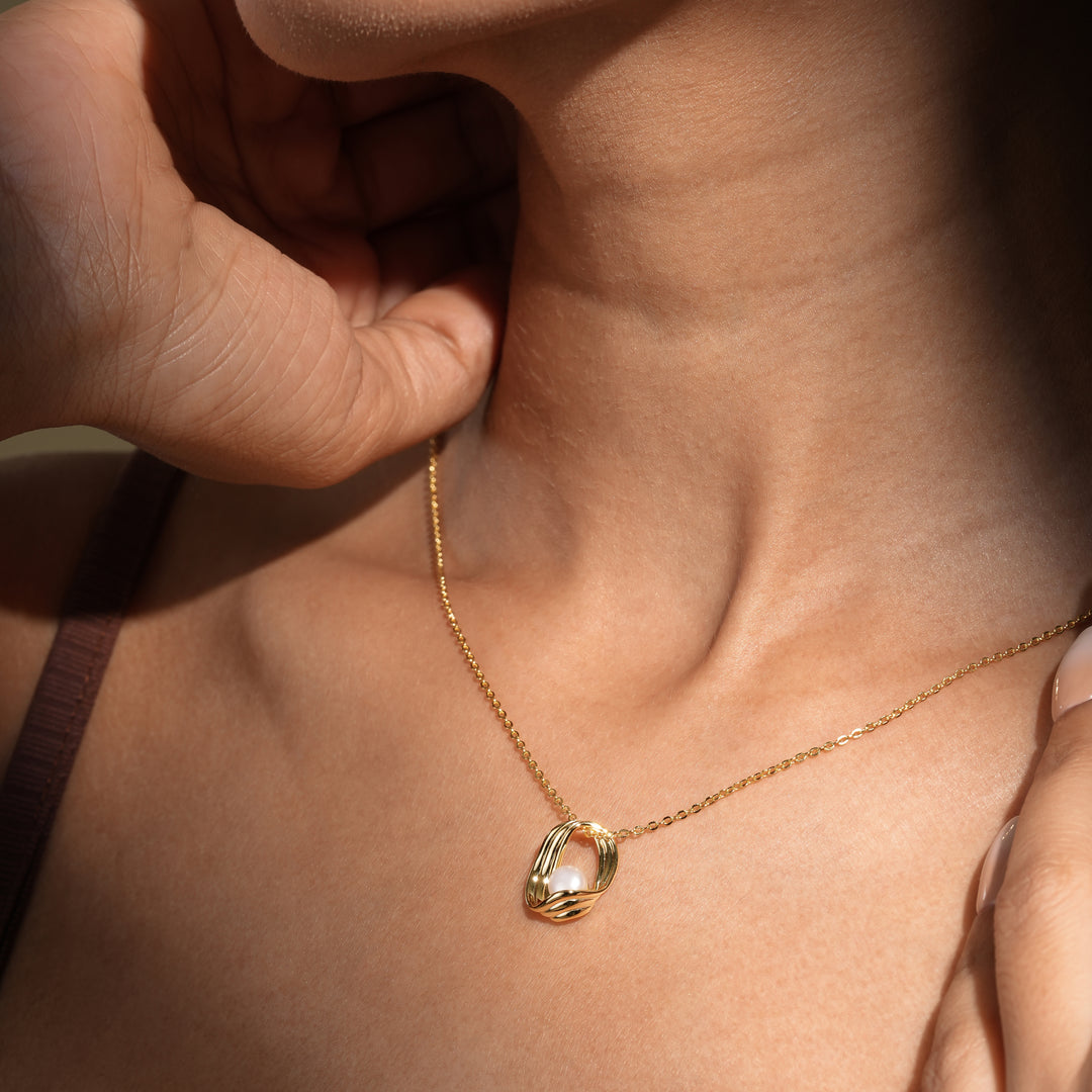 A close-up of a model wearing an elegant gold and pearl necklace from Zewory Jewellery. The modern, sculptural cage pendant with a nestled pearl is showcased in dramatic lighting, resting gracefully in the hollow of her throat.