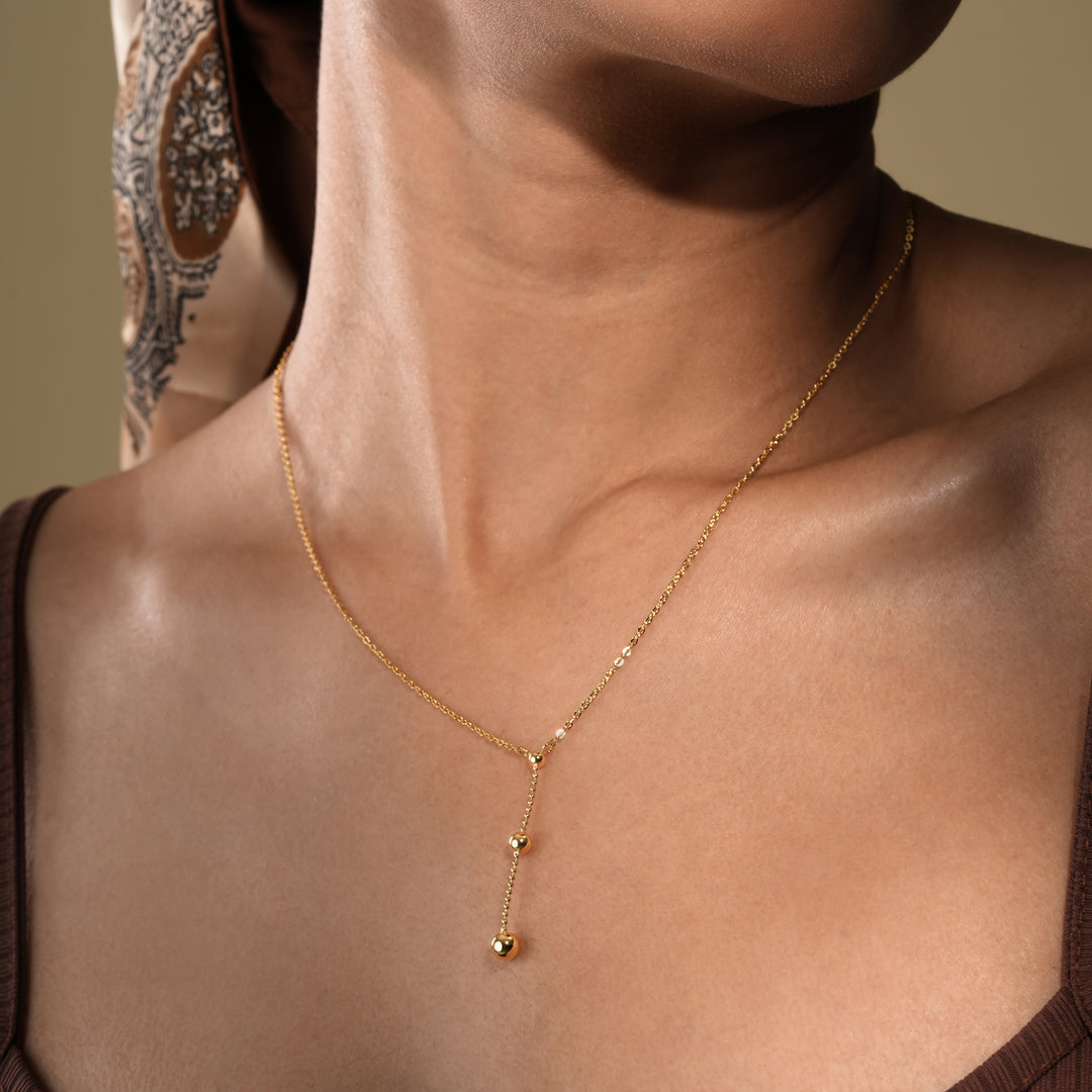 A close-up of a model wearing an elegant gold lariat necklace from Zewory Jewellery. The delicate Y-necklace features a drop chain with two polished gold beads that rest gracefully on her chest, showcased against her brown top.
