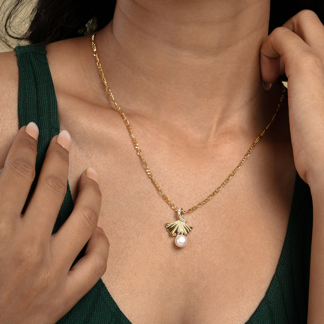 A close-up of a model wearing an elegant gold ginkgo leaf necklace from Zewory Jewellery. The detailed fan-shaped pendant, featuring a dangling pearl and a sparkling crystal accent, is showcased against her dark green top.