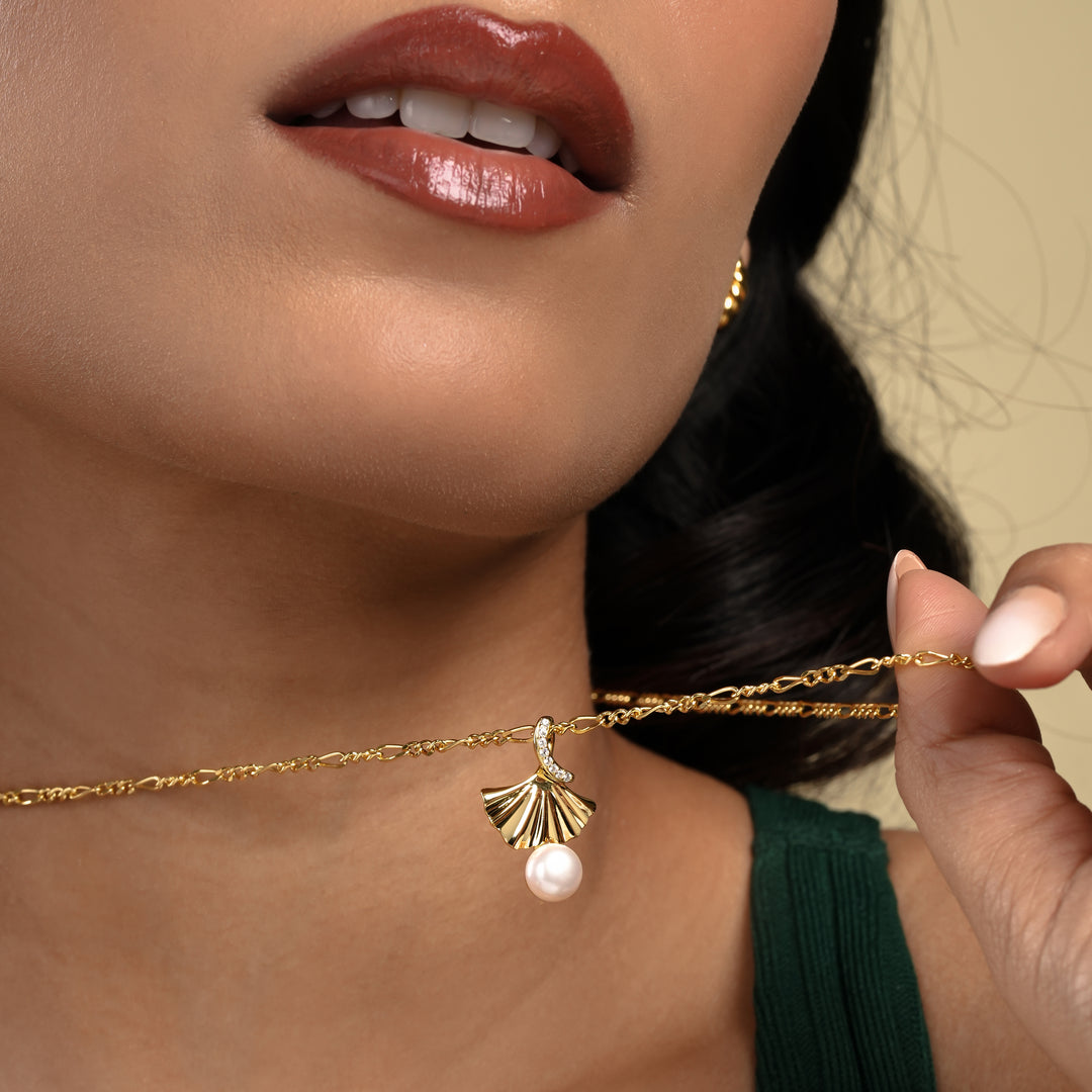 A close-up of a model wearing an elegant gold ginkgo leaf necklace from Zewory Jewellery. The detailed fan-shaped pendant, featuring a dangling pearl and a sparkling crystal accent, is showcased against her dark green top.