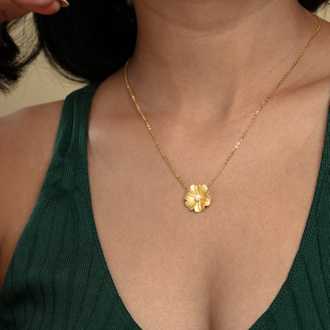 A close-up of a model's hands holding a delicate gold flower necklace from Zewory Jewellery. The detailed floral pendant, which features a matte gold finish and a central pearl, is elegantly displayed just below her chin.