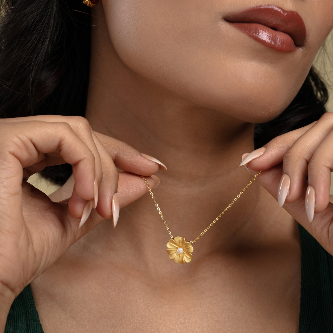 A close-up of a model's hands holding a delicate gold flower necklace from Zewory Jewellery. The detailed floral pendant, which features a matte gold finish and a central pearl, is elegantly displayed just below her chin.