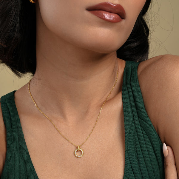 A close-up of a model wearing an elegant gold circle necklace from Zewory Jewellery. Her hands adjust the chain, showcasing the sparkling pavé crystal and twisted gold rope-textured pendant resting against her skin.
