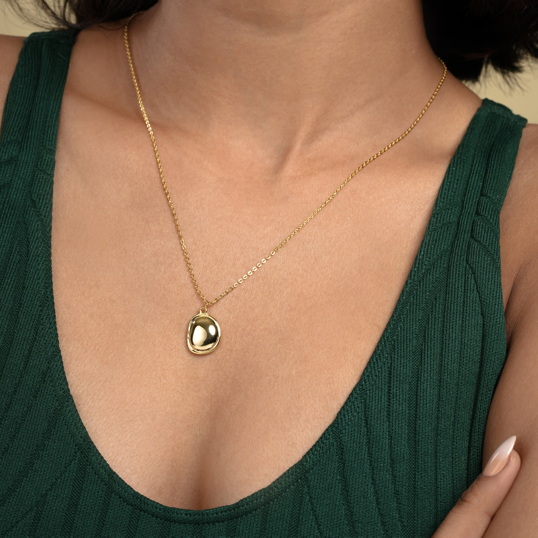 A close-up of a model wearing a minimalist gold necklace from Zewory Jewellery. The organic, pebble-shaped gemstone pendant with a smoky hue is showcased resting on her chest against a dark green top, perfect for everyday elegant style.