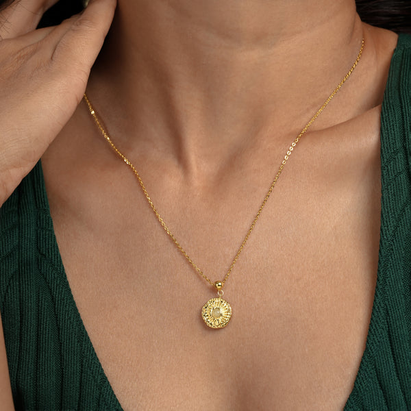 A close-up of a model wearing an elegant gold sunburst necklace from Zewory Jewellery. The detailed medallion pendant, featuring a central opalescent stone and radiating sun rays, rests on her chest against a dark green top.