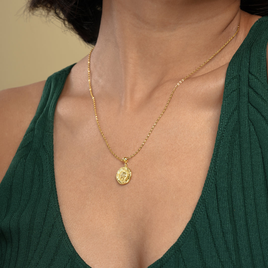 A close-up of a model wearing an elegant gold sunburst necklace from Zewory Jewellery. The detailed medallion pendant, featuring a central opalescent stone and radiating sun rays, rests on her chest against a dark green top.