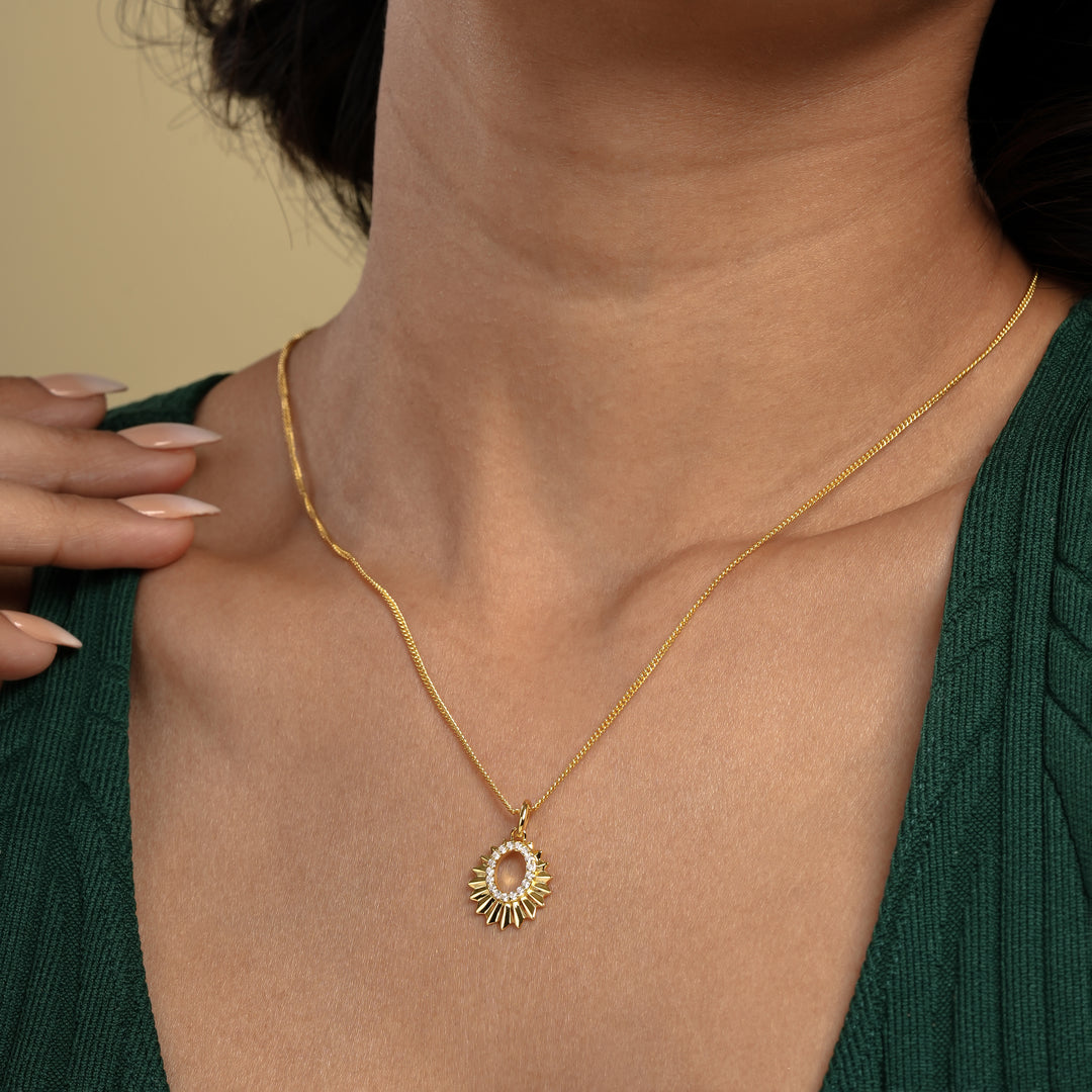 A close-up of a model wearing a radiant gold sun necklace from Zewory Jewellery. The celestial pendant, featuring faceted sun rays and a sparkling crystal-lined center, rests elegantly on her skin against her dark green top.