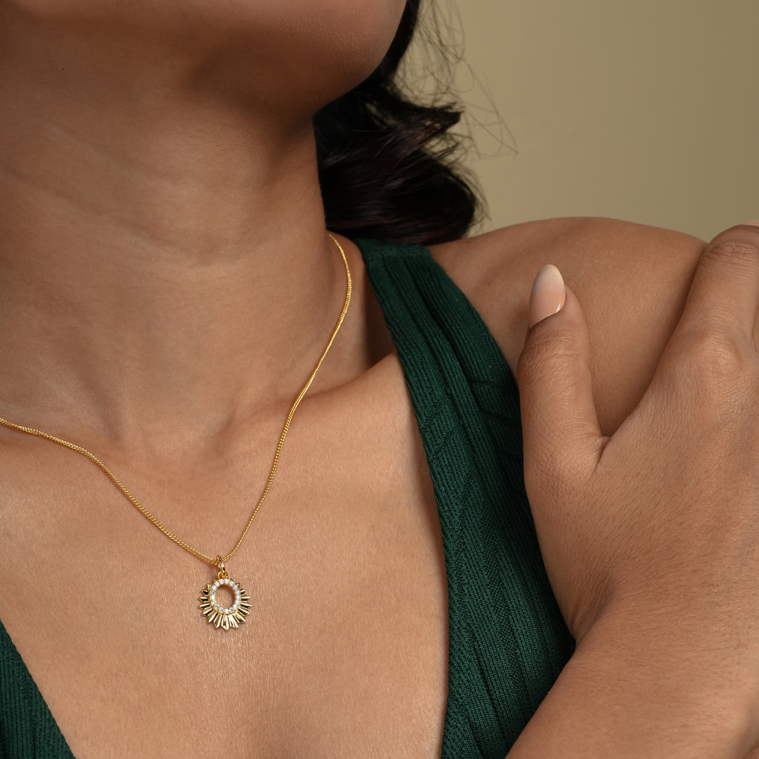 A close-up of a model wearing a radiant gold sun necklace from Zewory Jewellery. The celestial pendant, featuring faceted sun rays and a sparkling crystal-lined center, rests elegantly on her skin against her dark green top.