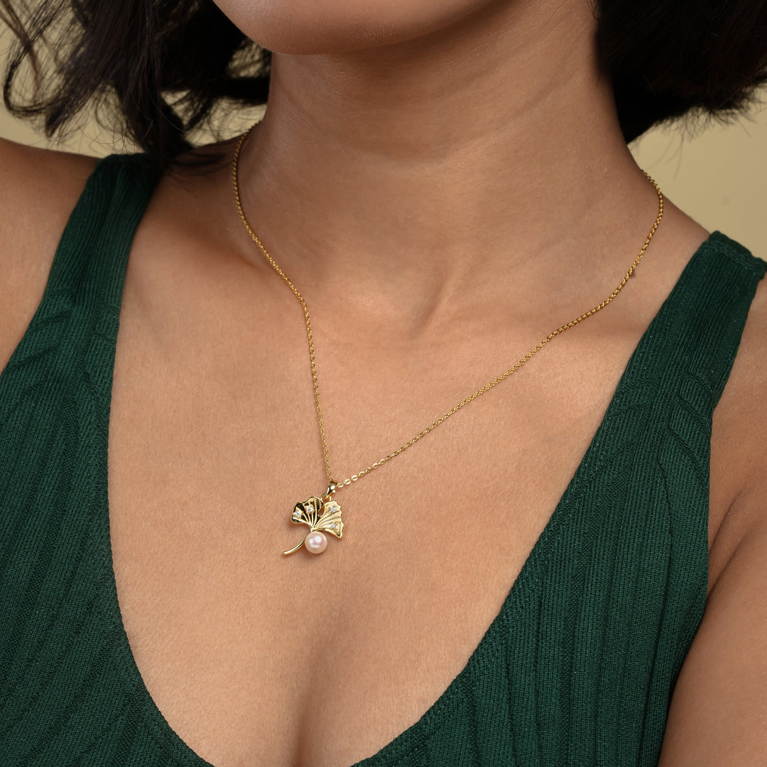 A close-up of a woman wearing a delicate gold necklace with a ginkgo leaf and pearl pendant against her skin. She is wearing a dark green ribbed top.