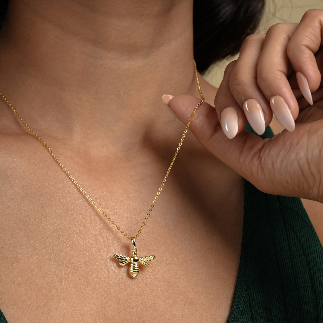 A close-up of a model wearing a delicate gold bee necklace from Zewory Jewellery. Her hand gently holds the chain, showcasing the detailed bumblebee pendant as it rests elegantly against her skin.
