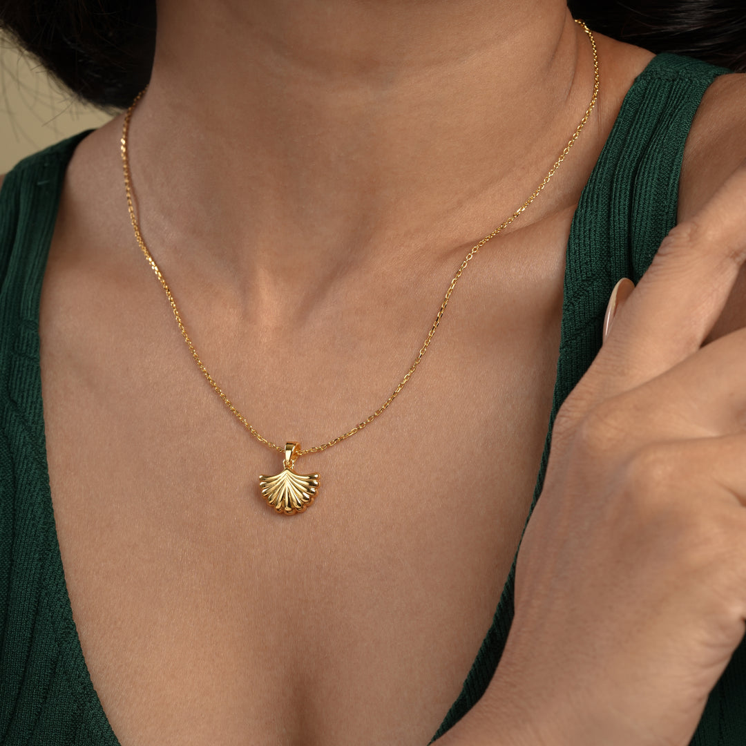 A close-up of a model wearing a delicate gold seashell necklace from Zewory Jewellery. The elegant, ribbed scallop shell pendant is showcased against her dark green top, perfect for a chic, ocean-inspired look.