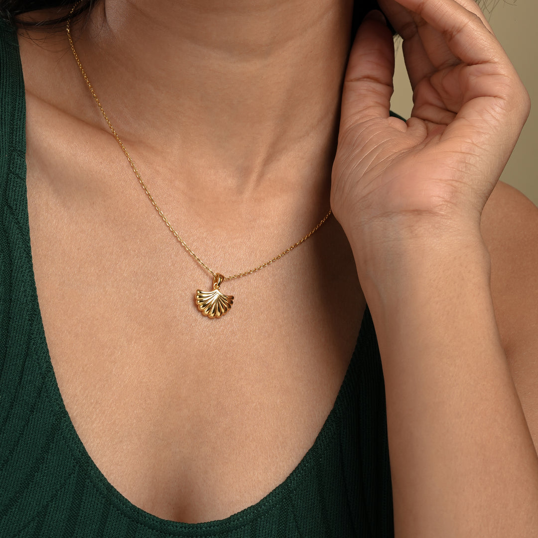 A close-up of a model wearing a delicate gold seashell necklace from Zewory Jewellery. The elegant, ribbed scallop shell pendant is showcased against her dark green top, perfect for a chic, ocean-inspired look.