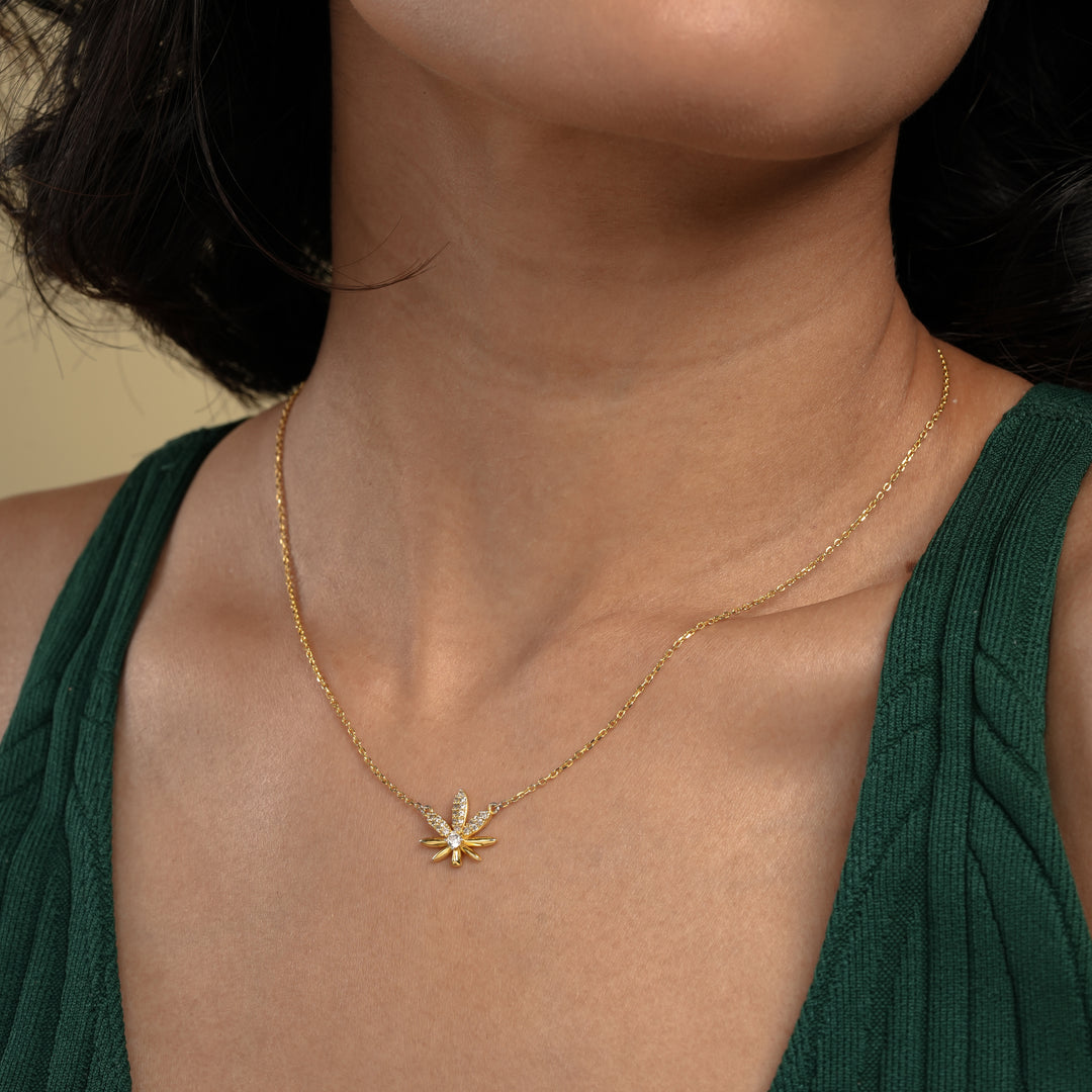 A close-up of a model wearing a delicate gold cannabis leaf necklace from Zewory Jewellery. The sparkling, crystal-encrusted pot leaf pendant rests elegantly against her skin, showcased with her dark green top.