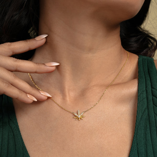 A close-up of a model wearing a delicate gold cannabis leaf necklace from Zewory Jewellery. The sparkling, crystal-encrusted pot leaf pendant rests elegantly against her skin, showcased with her dark green top.