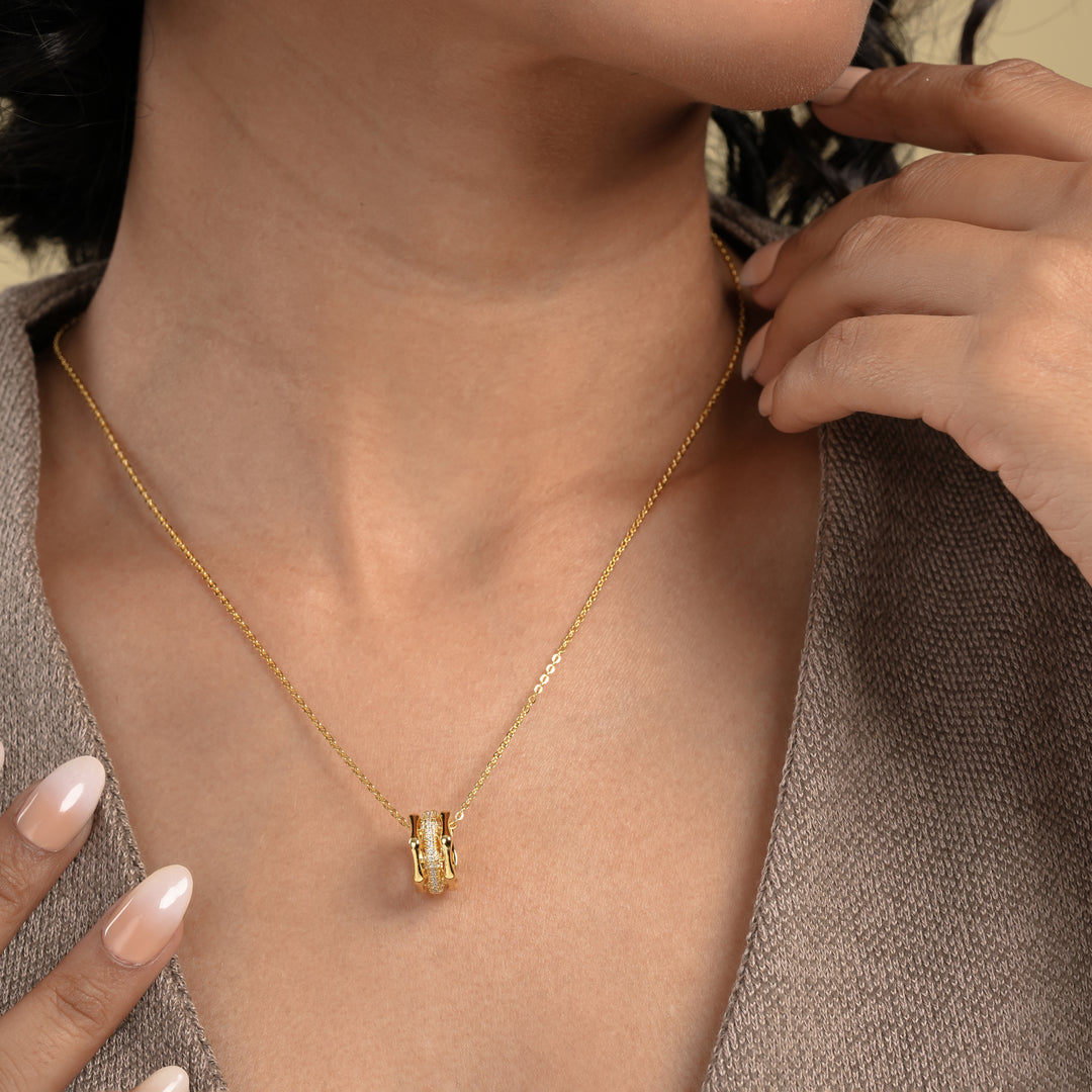 A close-up of a model wearing an elegant gold necklace from Zewory Jewellery, featuring a unique barrel pendant. The pendant's central pavé crystal band sparkles against her skin, showcasing a sophisticated piece of fashion jewellery.