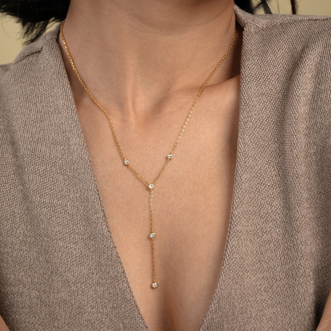 A close-up of a model wearing an elegant gold lariat necklace from Zewory Jewellery. The delicate Y-necklace, featuring five sparkling bezel-set crystals, is showcased resting gracefully against her skin and a neutral-coloured top.