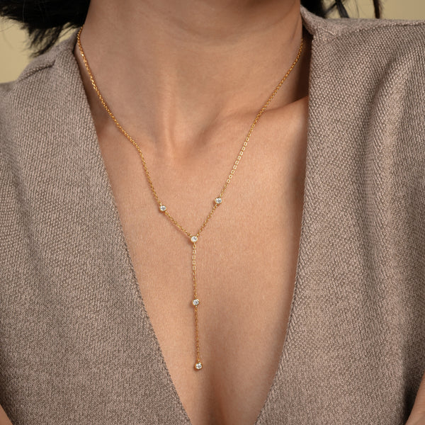 A close-up of a model wearing an elegant gold lariat necklace from Zewory Jewellery. The delicate Y-necklace, featuring five sparkling bezel-set crystals, is showcased resting gracefully against her skin and a neutral-coloured top.