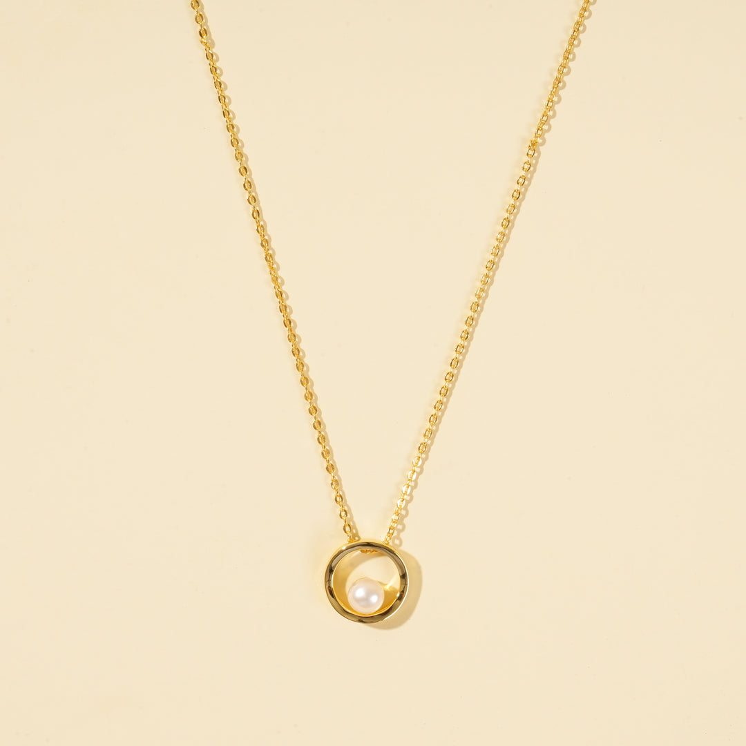 A clean, minimalist product shot of a gold and pearl necklace on a neutral cream background. The image clearly displays the modern design of a single, lustrous pearl held within a polished gold circle pendant.