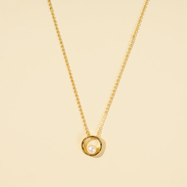 A clean, minimalist product shot of a gold and pearl necklace on a neutral cream background. The image clearly displays the modern design of a single, lustrous pearl held within a polished gold circle pendant.