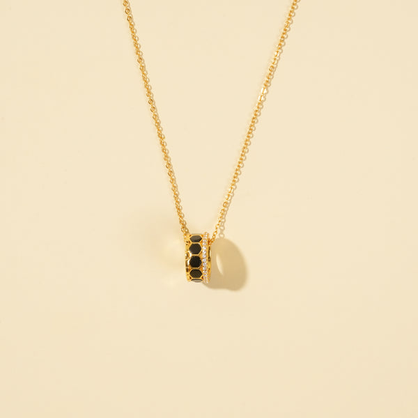 A clean, minimalist product shot of a gold honeycomb necklace on a neutral cream background. The image clearly displays the intricate barrel pendant, which features black hexagonal inlays and two bands of sparkling pavé crystals.