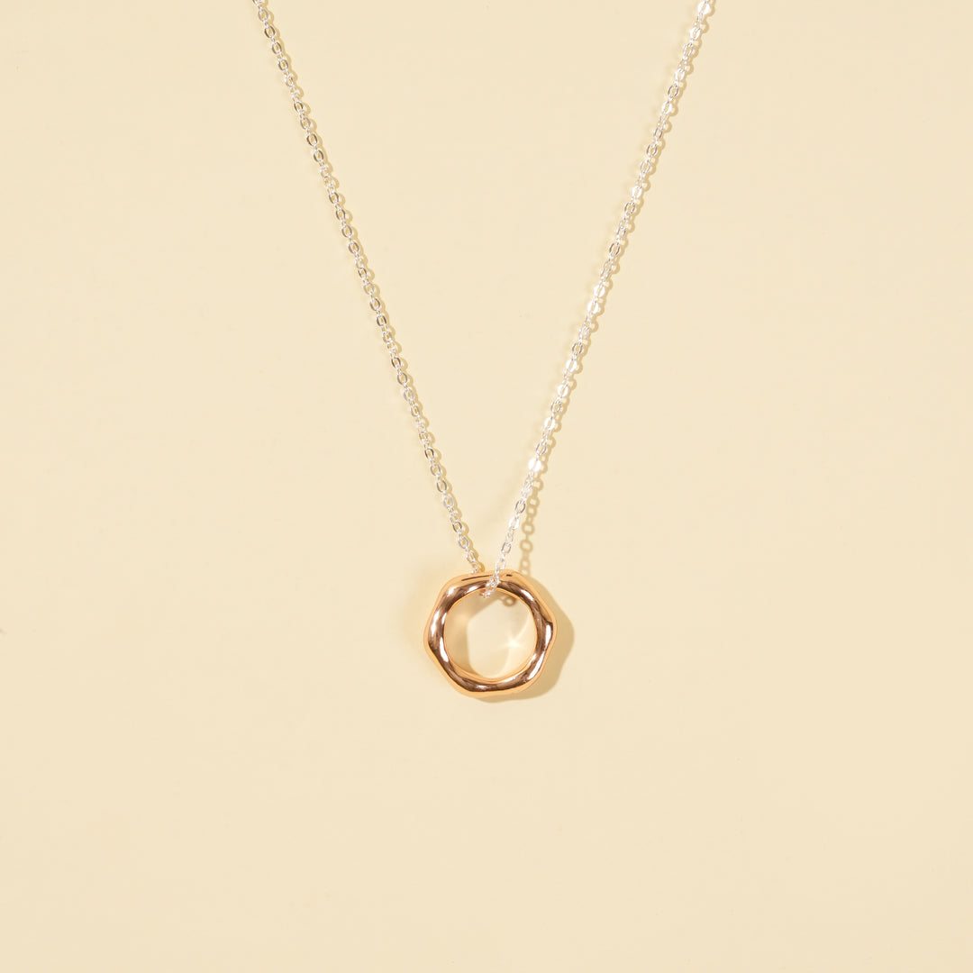 A clean, minimalist product shot of a two-tone necklace on a neutral cream background. The image clearly displays the delicate silver chain and the modern, organic-shaped rose gold open circle pendant.